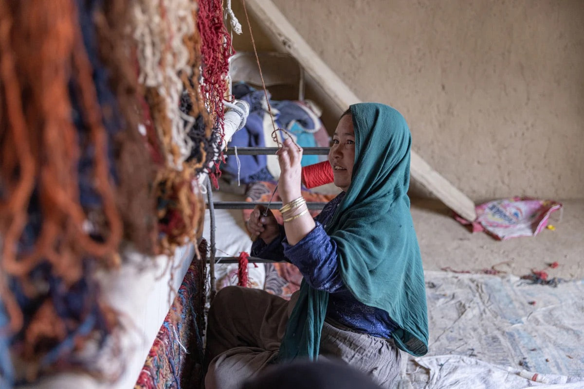 Woman weaving a rug on a vertical loom beside bundles of yarn