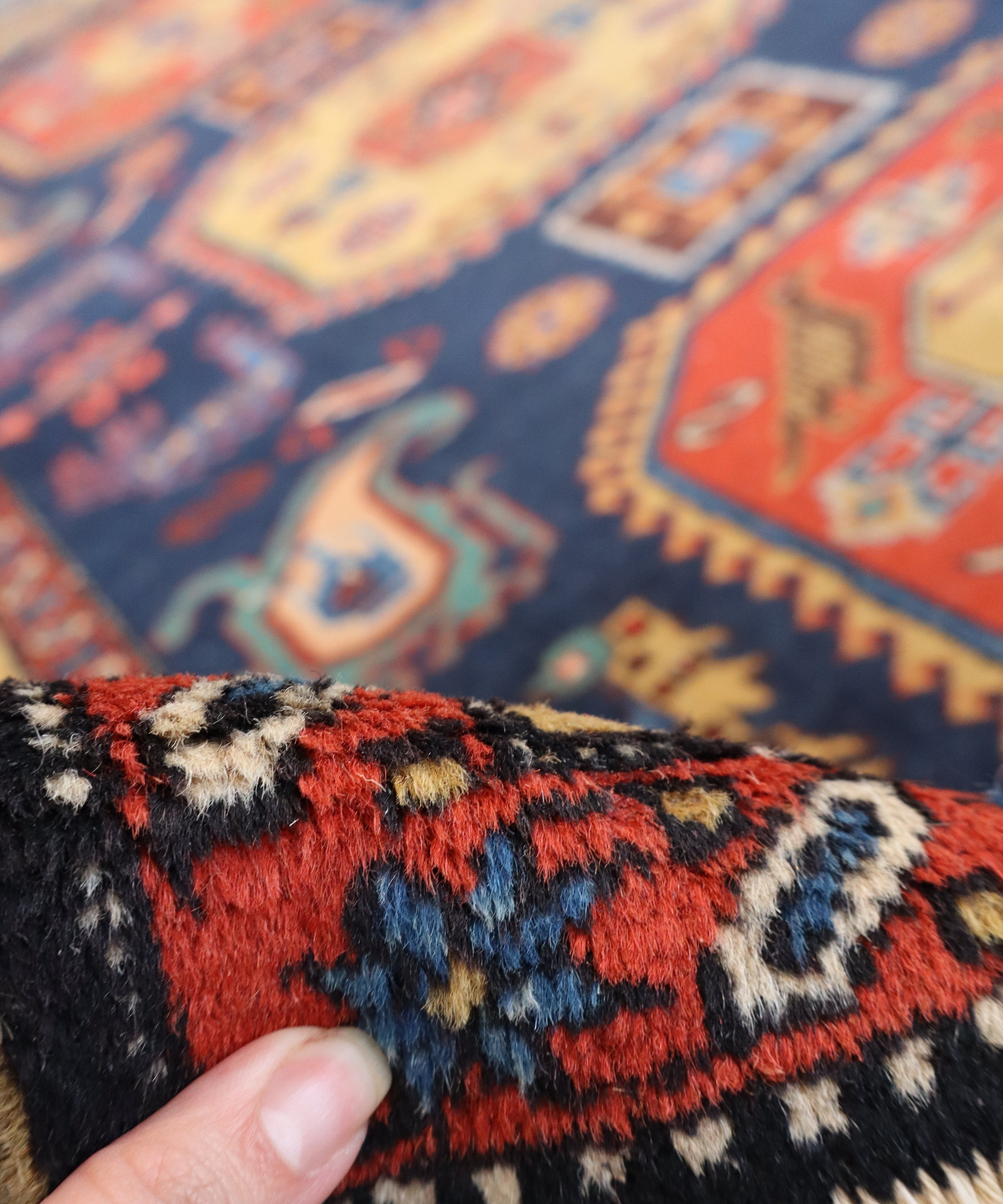 Persian mesh rug corner with multicolor pattern and frayed pile, fingertip visible