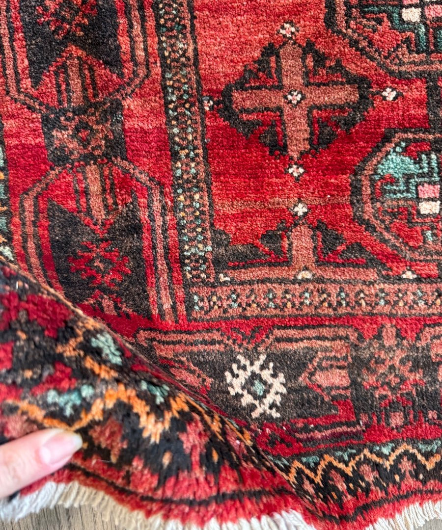 Red Persian Baluch rug close-up with black and teal geometric motif and fringe.