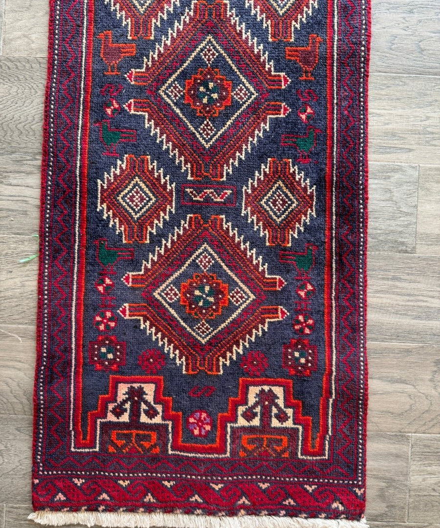 Persian Baluch runner rug with dark navy field, red border, diamond geometric motifs, fringe ends.