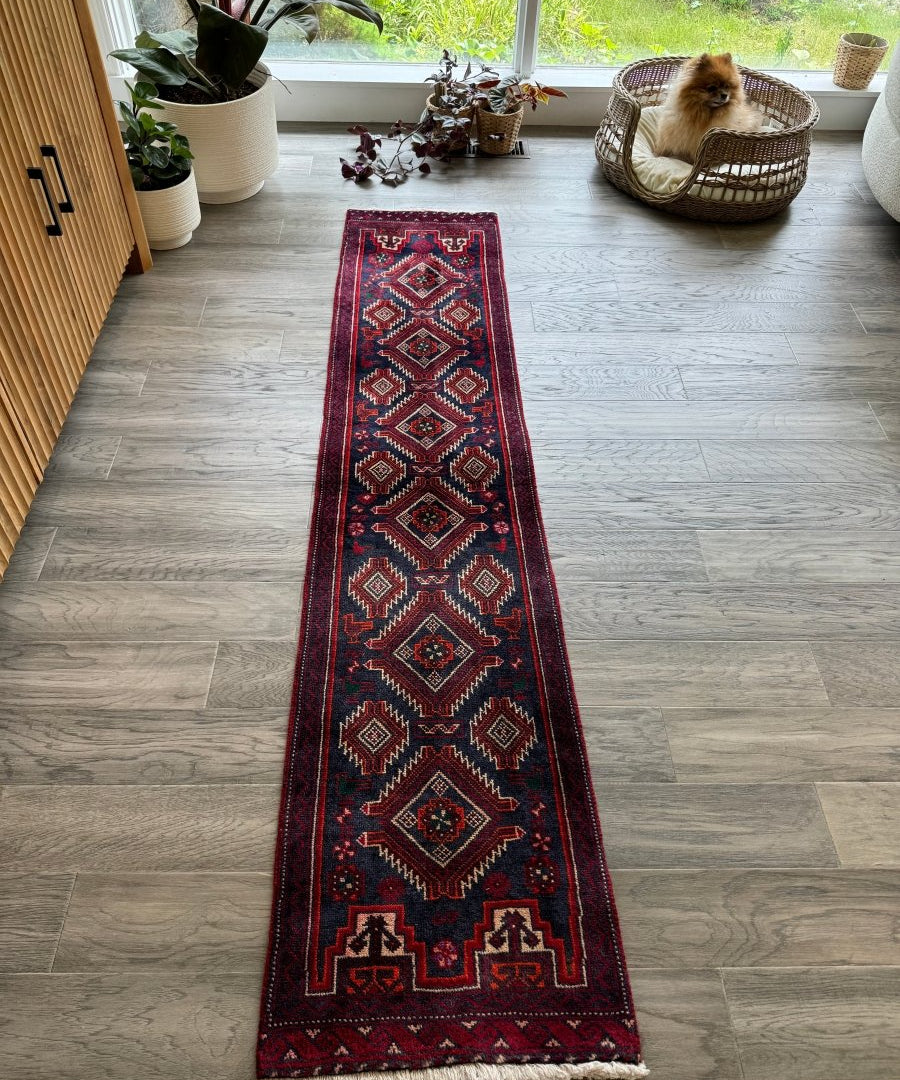 Narrow Persian Baluch runner rug, dark navy with red border and repeating diamond patterns, fringed.