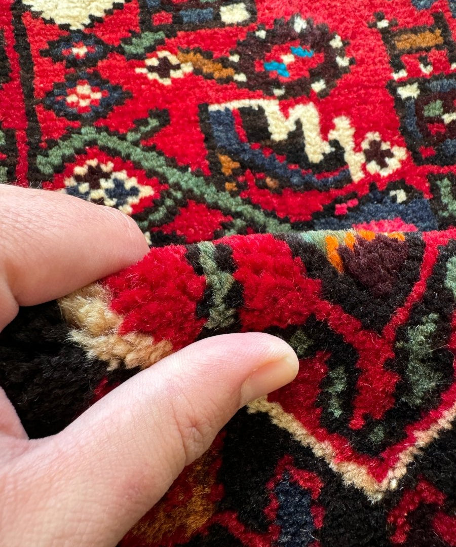 Rug pile detail in red and dark colors with geometric pattern; fingers gently touch.