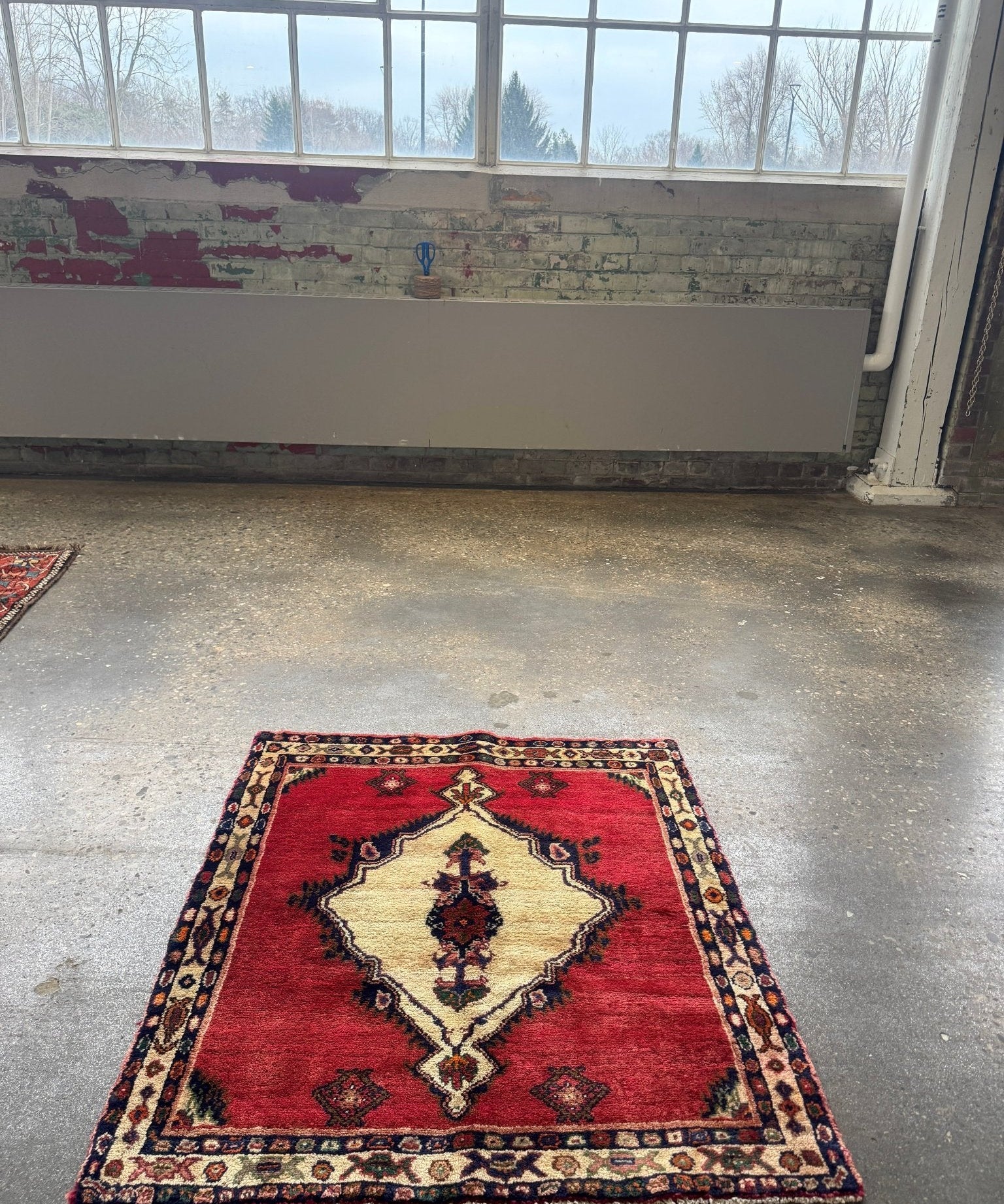 Small Persian Hamadan-style rug with red field and cream diamond medallion on gray floor.