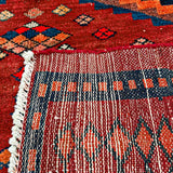 Red rug close-up showing navy border, orange diamond pattern, and striped fringe trim.
