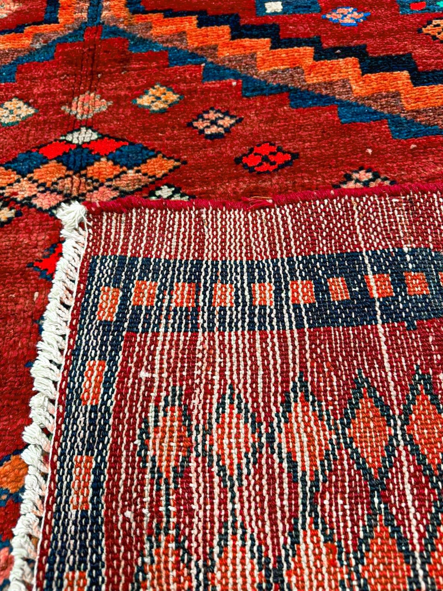 Red rug close-up showing navy border, orange diamond pattern, and striped fringe trim.