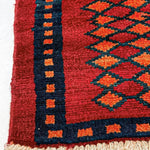 Red rug corner with navy border and repeating diamond motifs; cream short fringe edge.