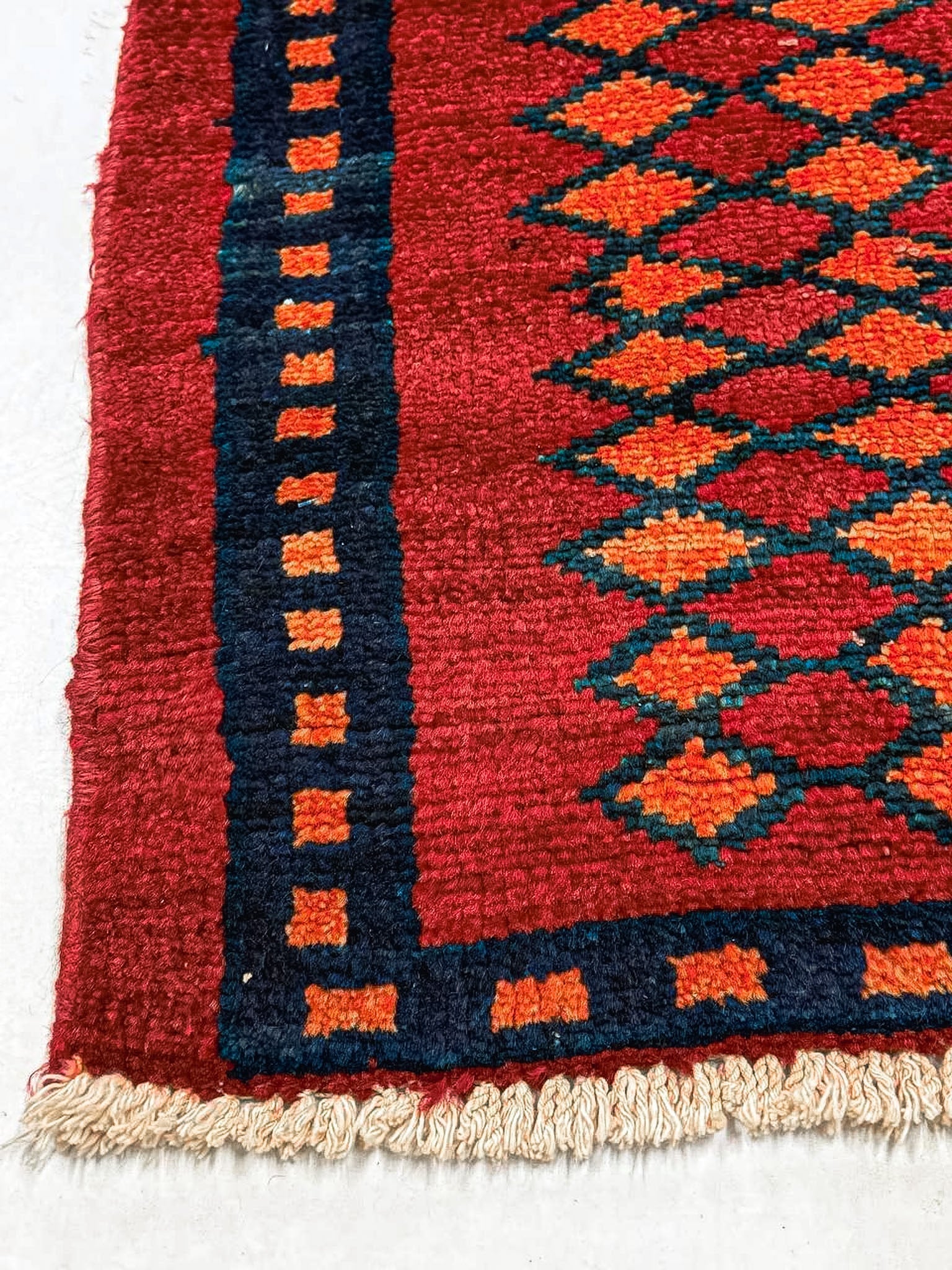 Red rug corner with navy border and repeating diamond motifs; cream short fringe edge.