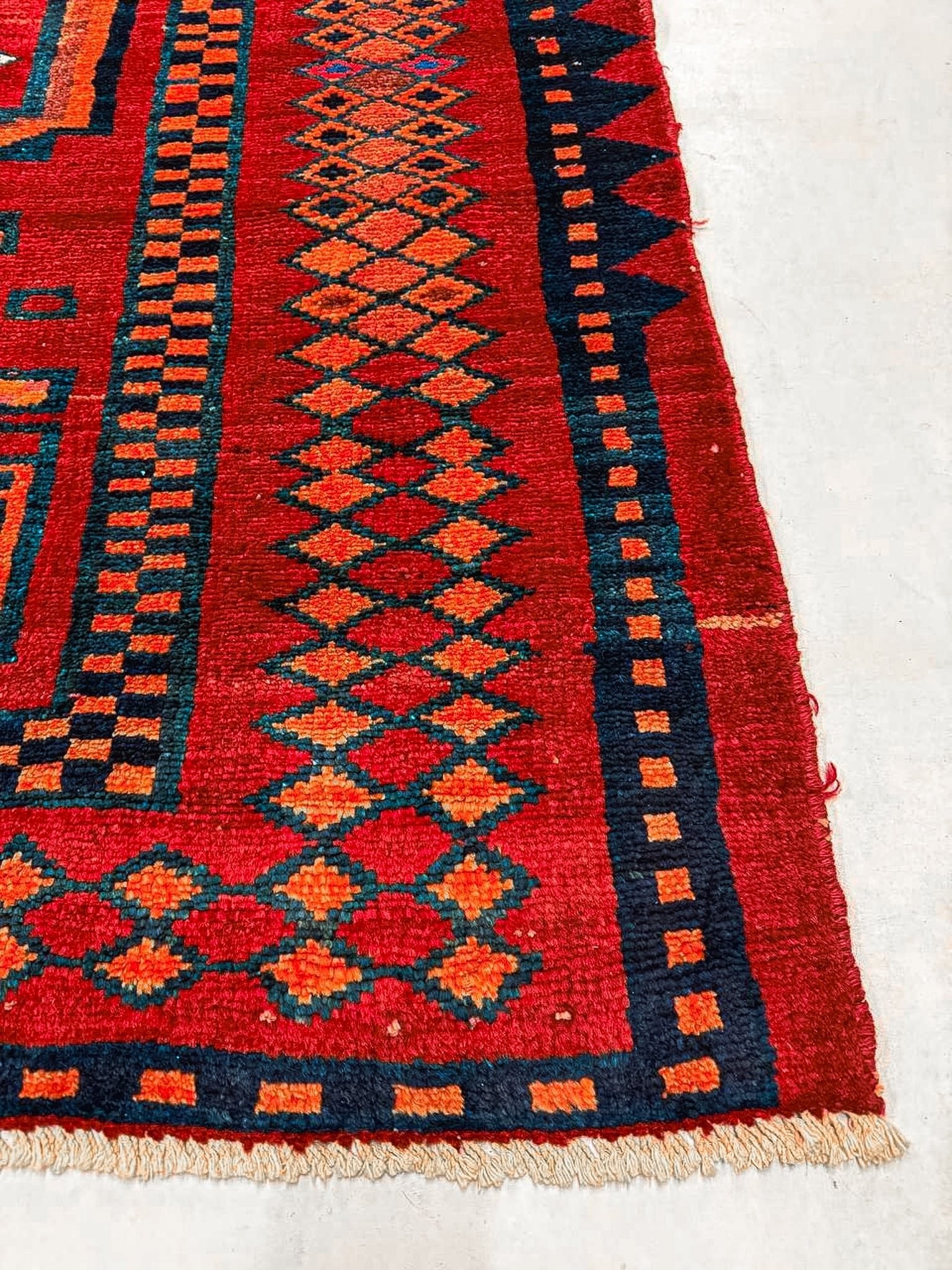 Red rug with navy and teal geometric motifs; orange accents; visible cream fringe at edge.