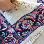 Hand holding rug corner; off-white pile with navy-red ornate border and fringe