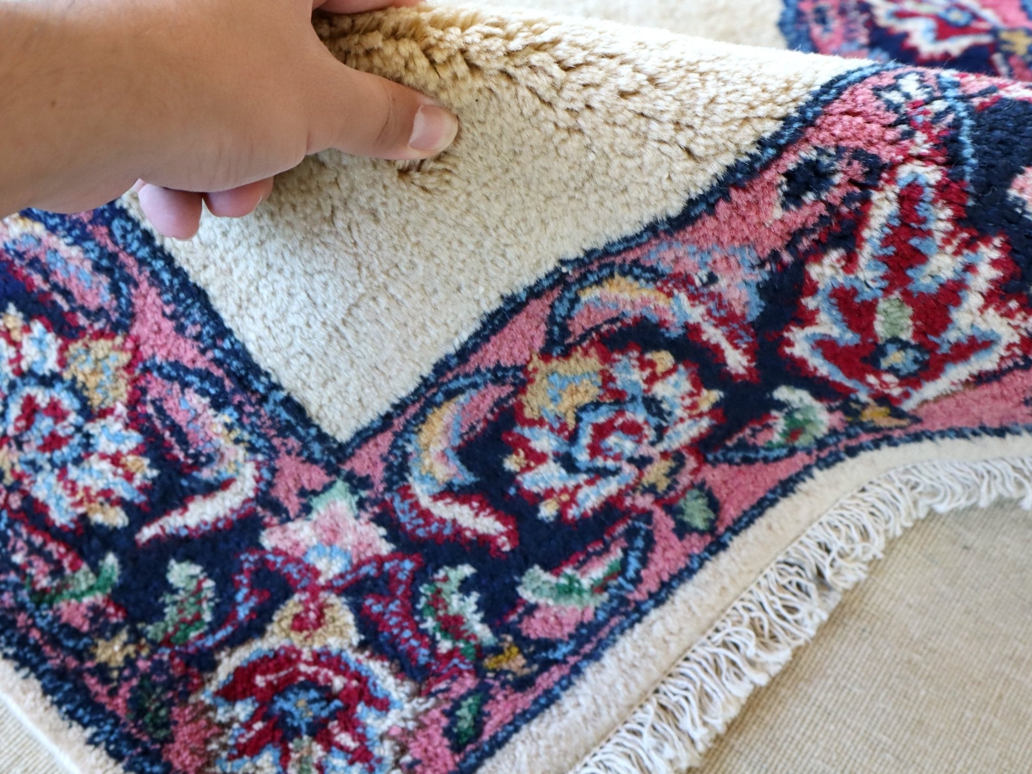 Hand holding rug corner; off-white pile with navy-red ornate border and fringe