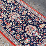 Persian Qom runner rug corner view: navy floral vines, multicolor rosettes, red border