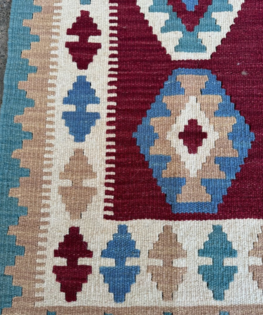 Geometric kilim rug section with blue and beige motifs on maroon, showing flat weave lines.