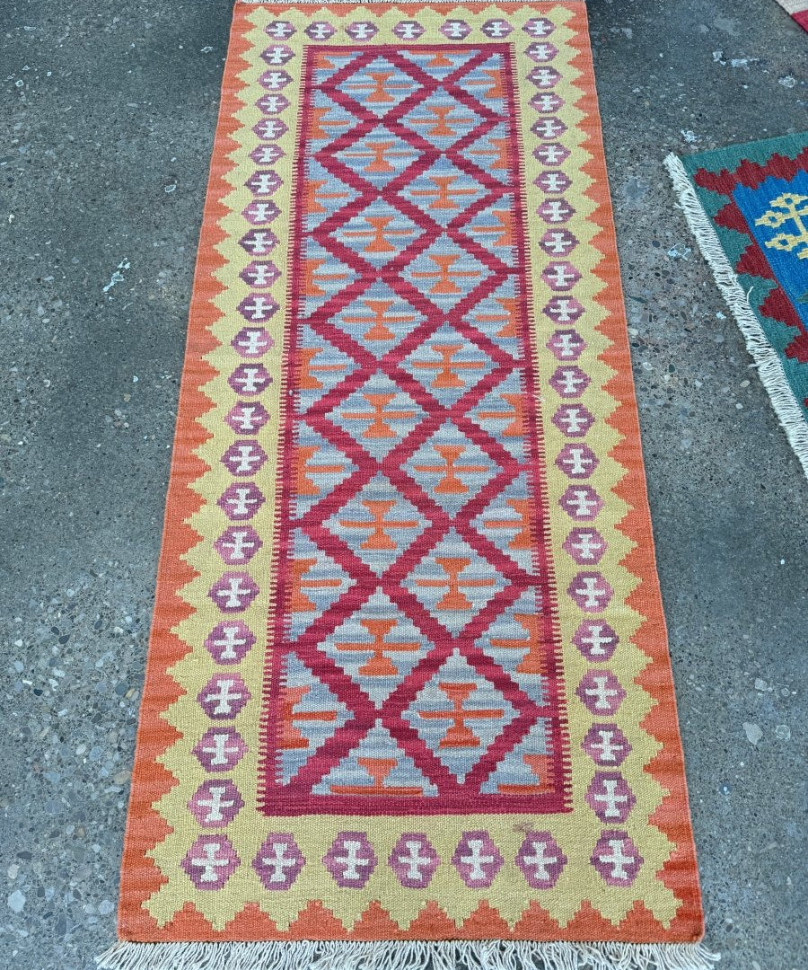Orange runner rug with geometric diamond pattern in pink, blue, and purple; fringed ends.