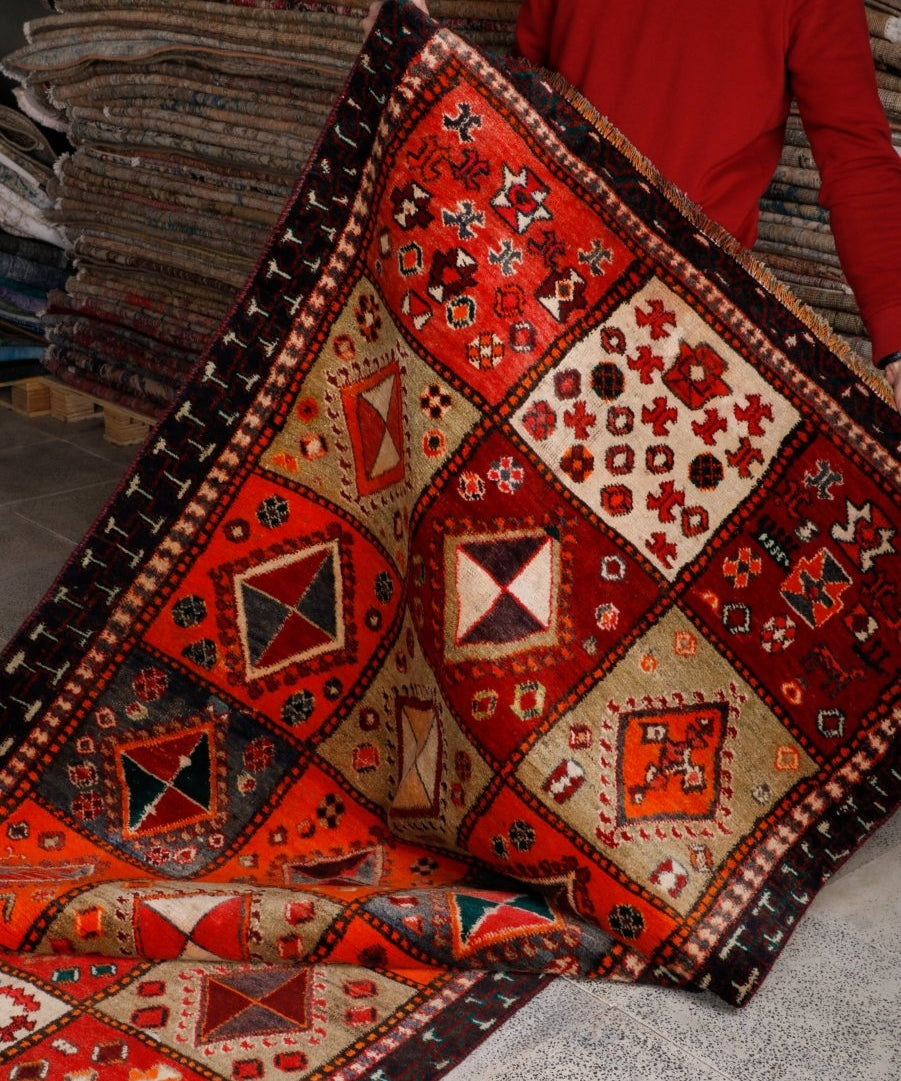 Persian Shiraz rug draped on floor stacks; red-orange field with cream motifs and dark trim.
