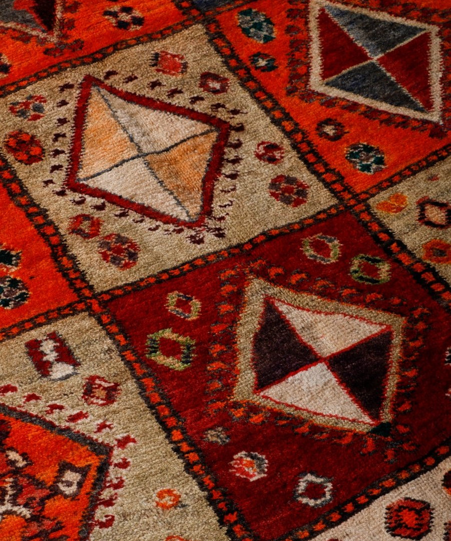 Close-up of Persian Shiraz rug: red-orange, cream, and dark geometric pattern on textured pile.