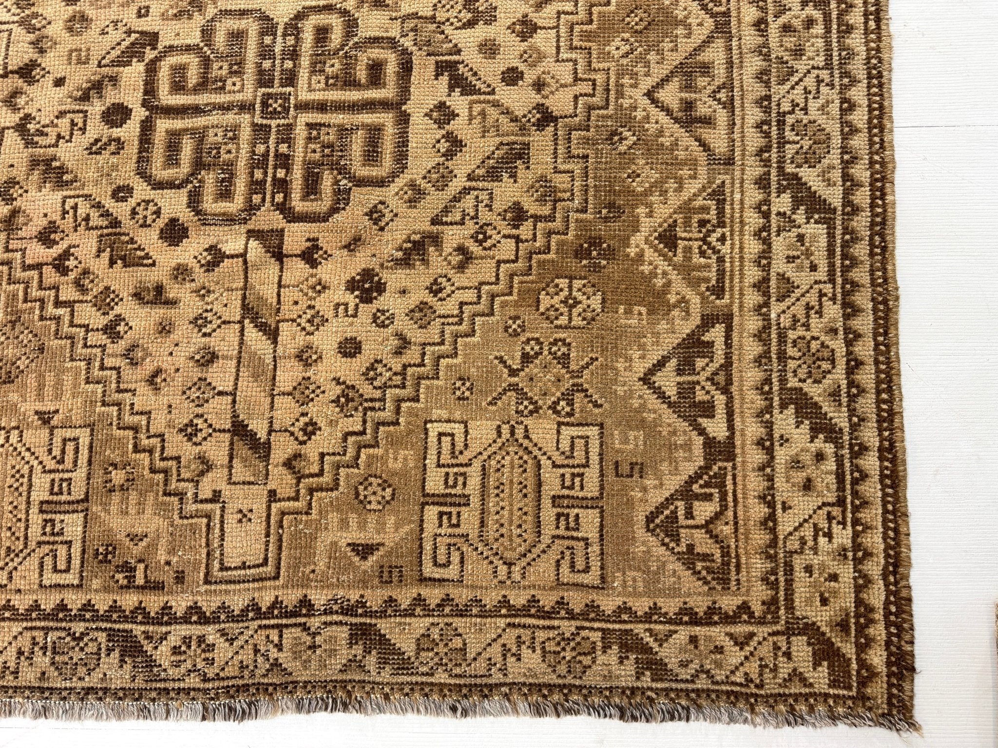 Persian rug corner with tan and dark brown patterns, narrow stripes, and short fringe edge.