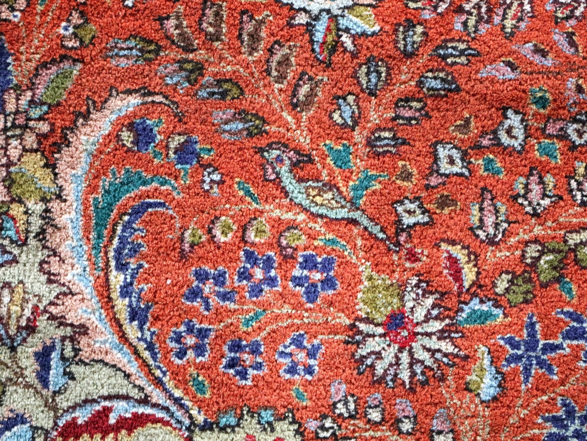 Rug close-up with red-orange ground, blue floral motifs, and teal accent details.