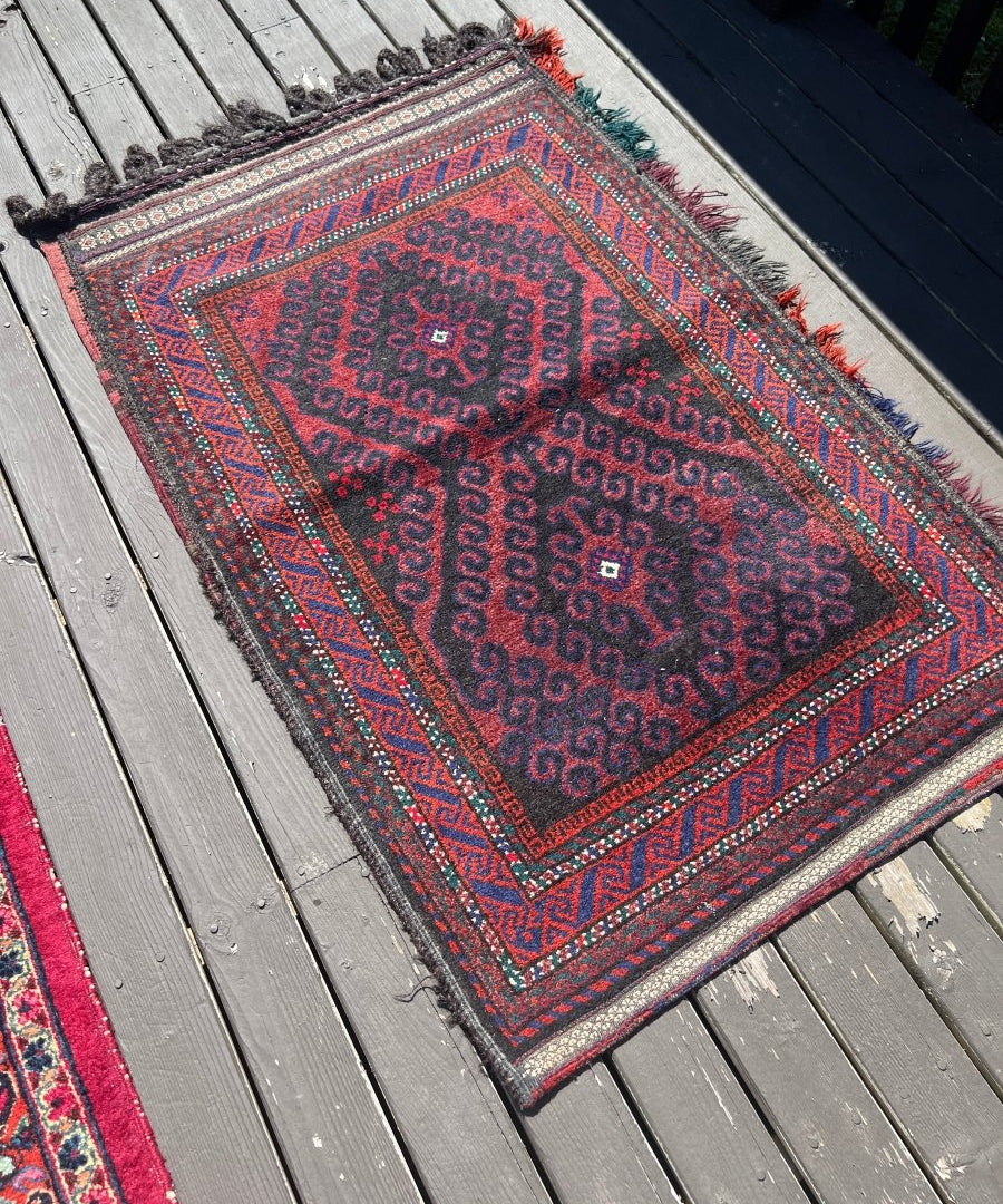 Antique Baluch saddlebag rug with red center and navy geometric pattern, fringed edges.