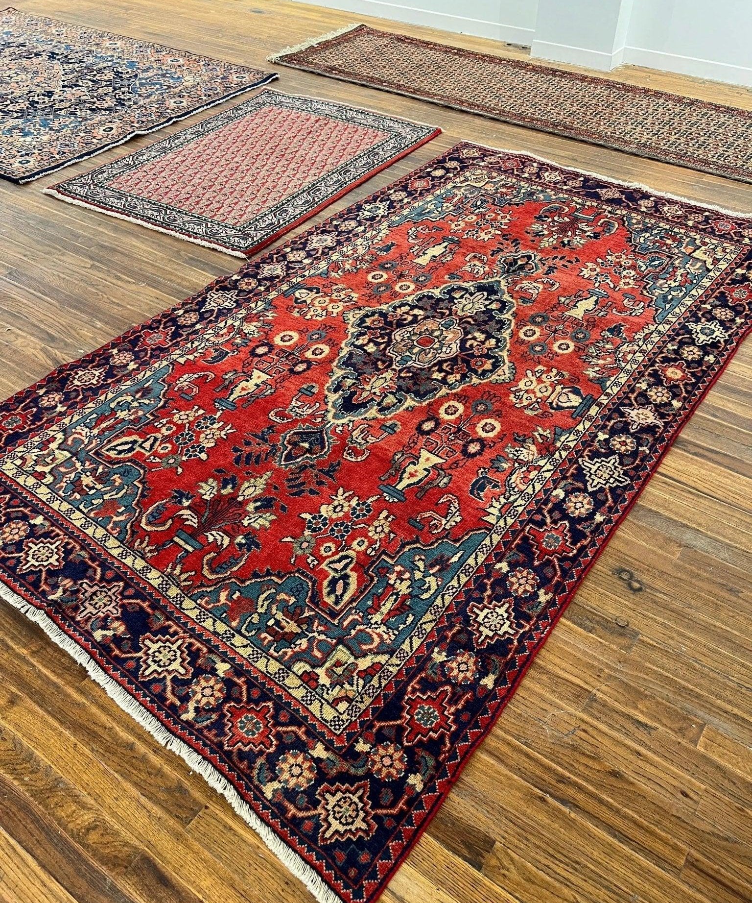 Red Persian-style rug on hardwood floor, ornate pattern and navy border with trimmed fringe.