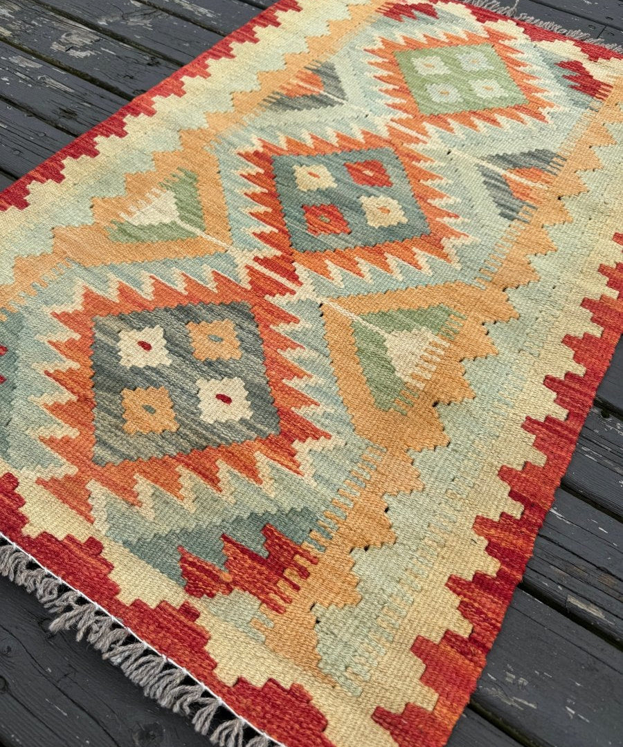 Kilim rug angled view with diamond pattern, red and cream border, teal and orange geometric blocks, fringe