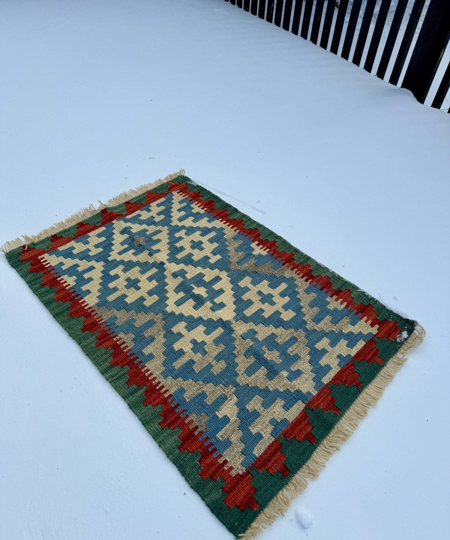 Small kilim rug angled view with blue pattern, red and green border, and cream fringe